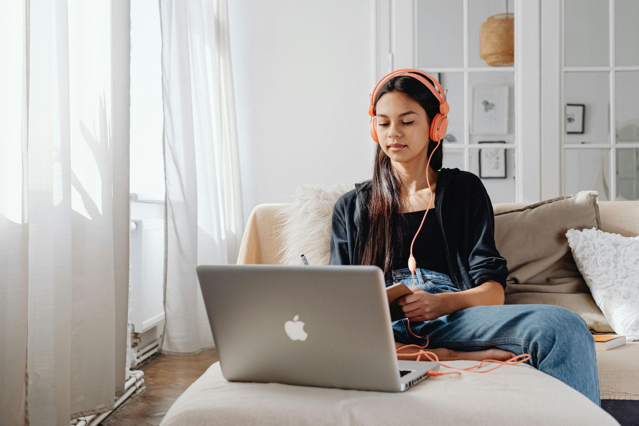 Girl with headphones taking notes while watching an online class on her laptop.